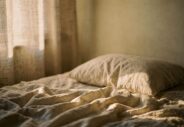 Lit défait et draps de lin clairs éclairés par la première lumière du jour à travers un rideau.webp