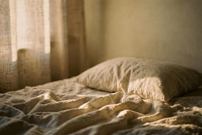 Lit défait et draps de lin clairs éclairés par la première lumière du jour à travers un rideau